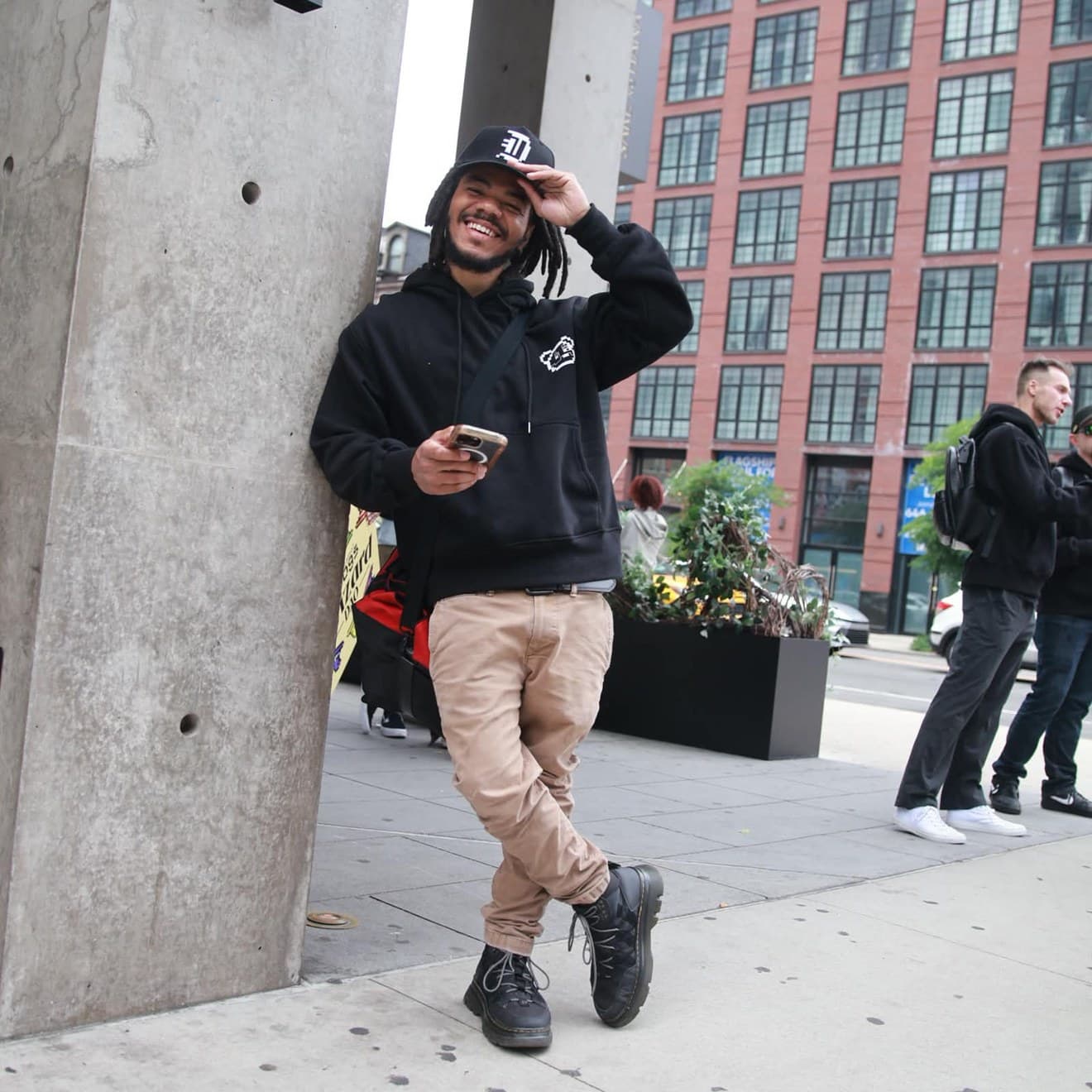 Corn (@CornKry) smiling on a city sidewalk, hand on his cap